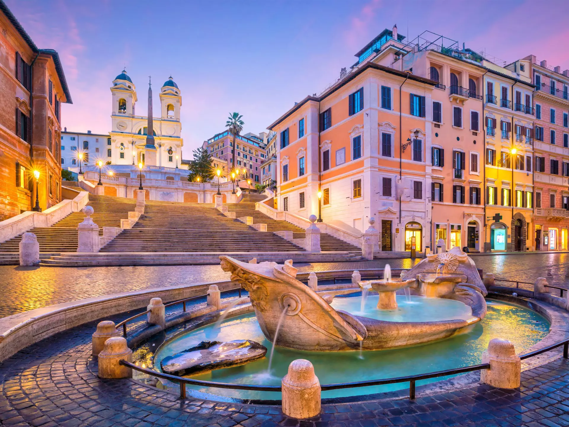 A series of steps lead down to a Roman plaza with cobbled stone streets and in the center of the photo is a marble foundation in the shape of a boat.