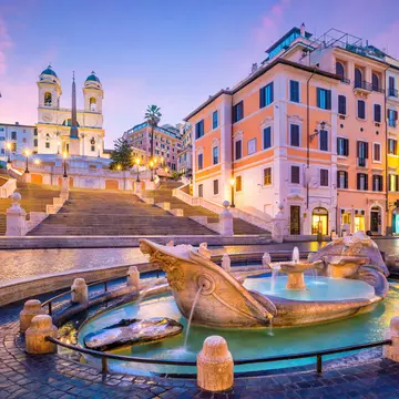A series of steps lead down to a Roman plaza with cobbled stone streets and in the center of the photo is a marble foundation in the shape of a boat.