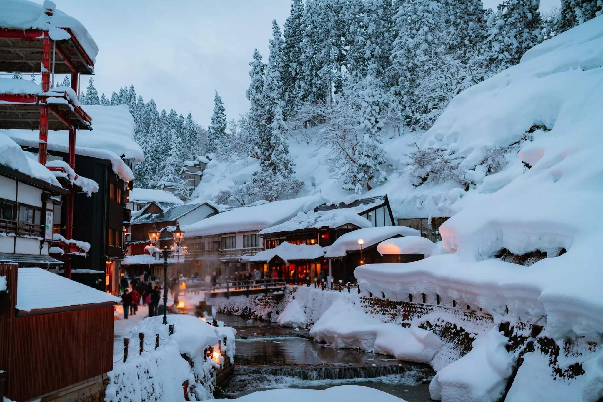 An onsen (hot springs) complex is pictured on a winter day. Heavy snow covers the roofs, and pine trees on a slope that rises behind.