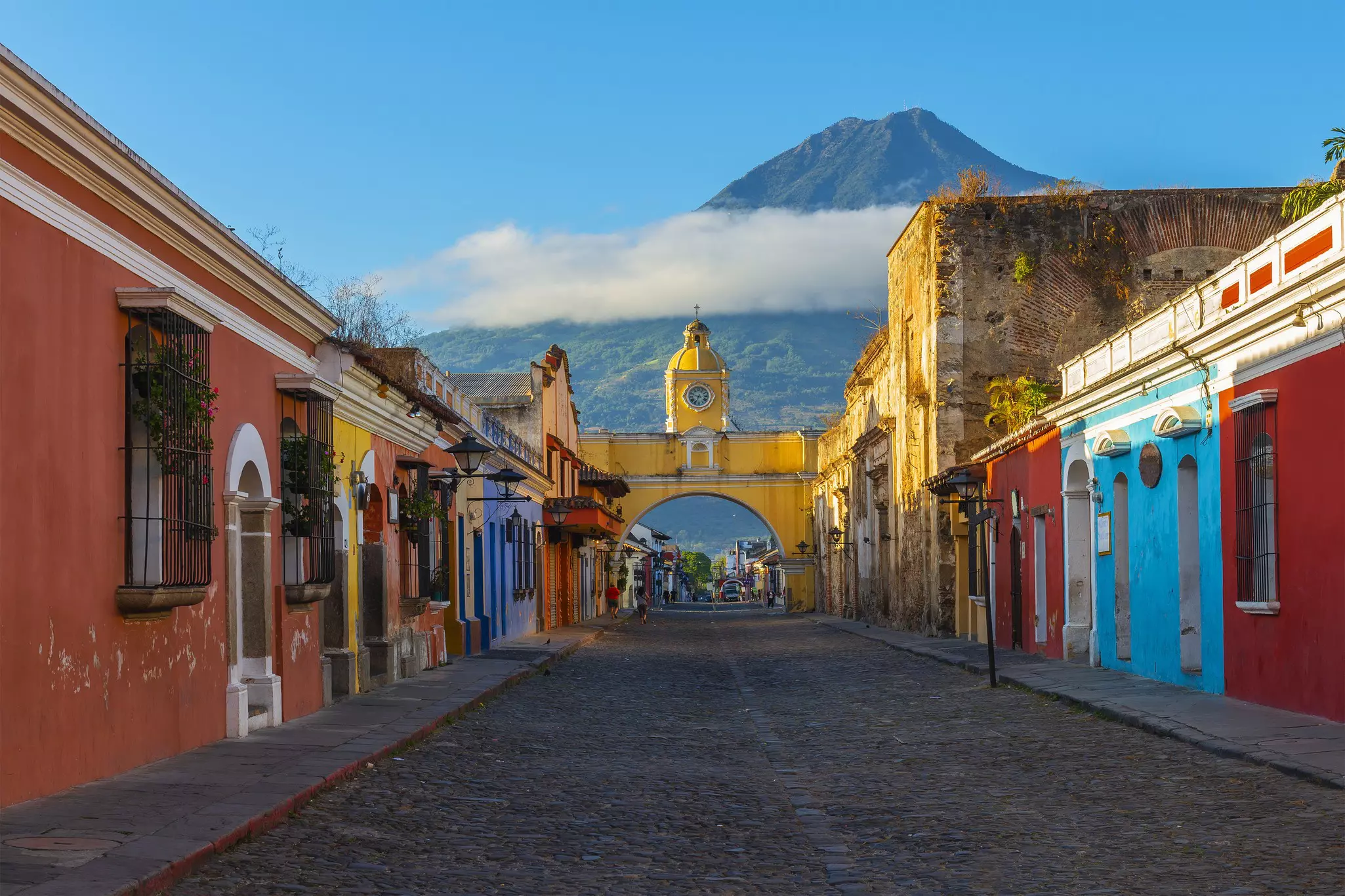 A cobbled street lined with colorful painted low-rise properties. A yellow arch with a clock on the top rises over the street. A large mountain covered with cloud looms over the town