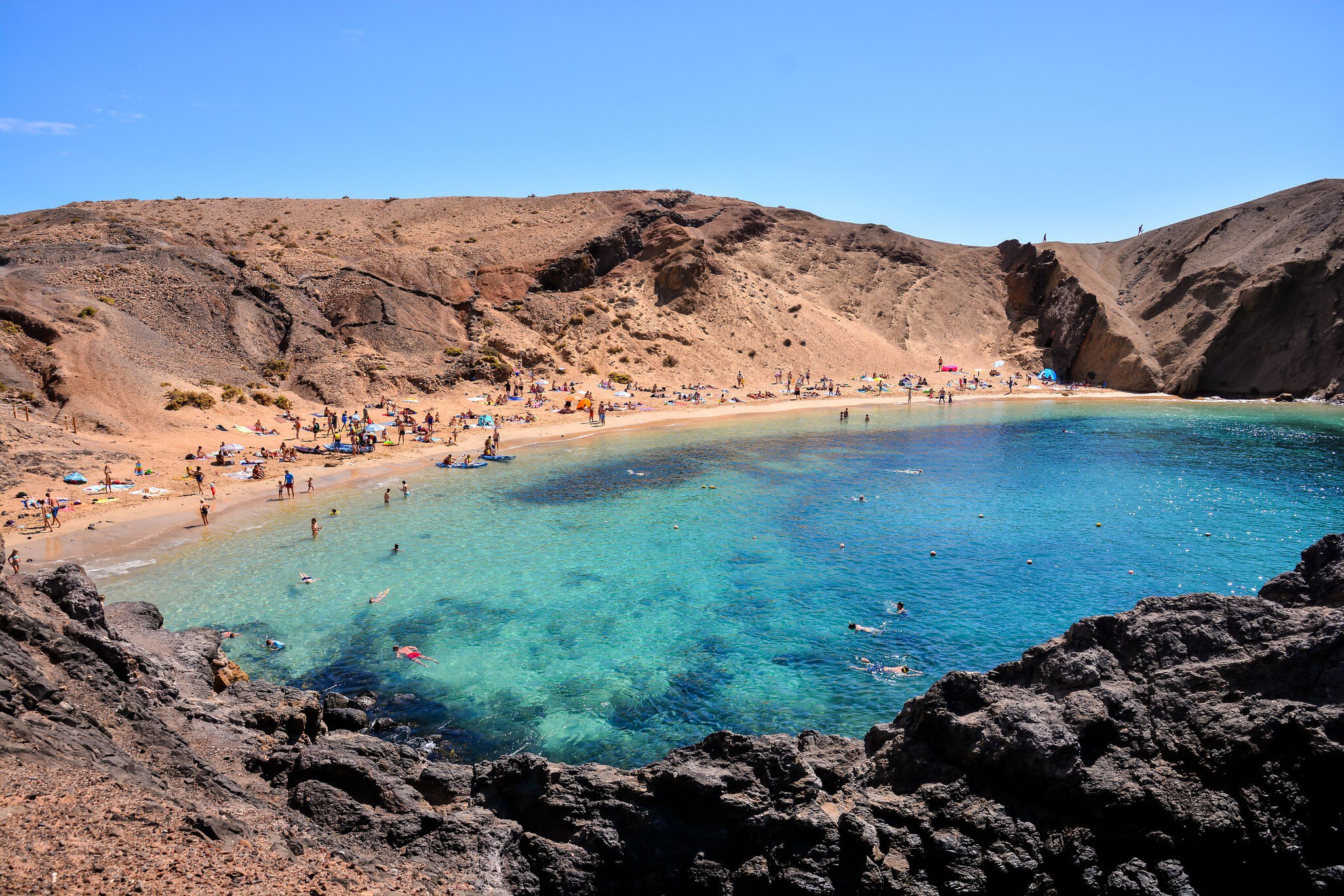 Crescent of golden sand crowded with beachgoers below brown volcanic slopes, dark rocks in the foreground