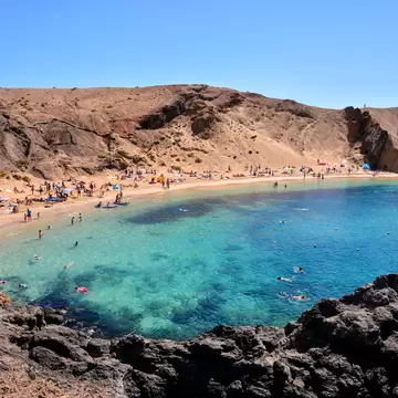 Crescent of golden sand crowded with beachgoers below brown volcanic slopes, dark rocks in the foreground