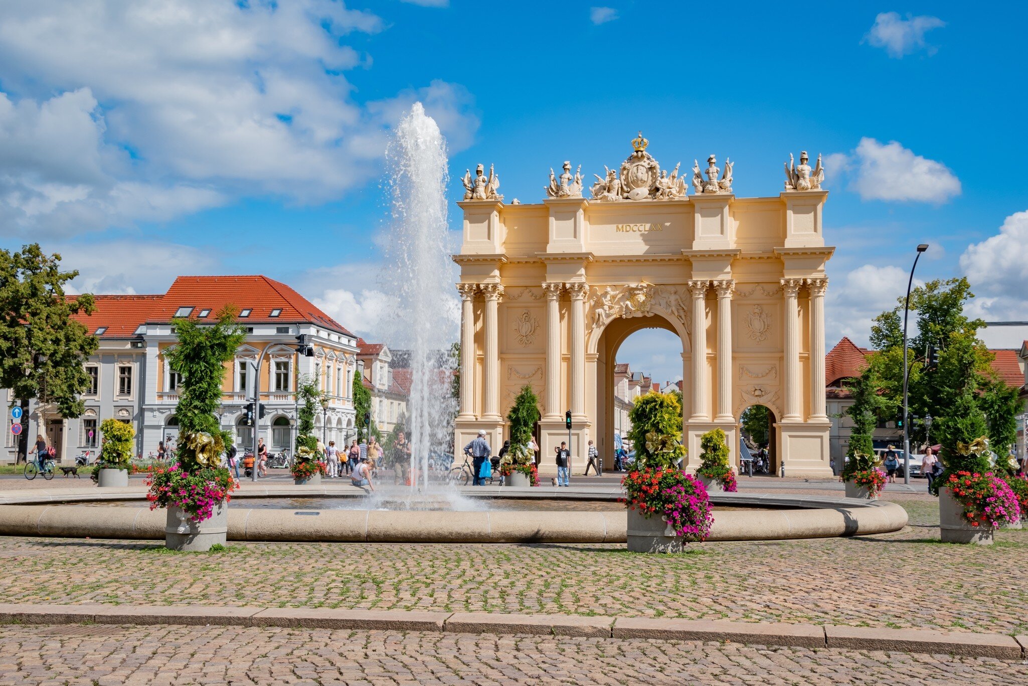 Potsdam's yellow Brandenburg Gate arch inscribed MDCCLXX, with a fountain and flower planters on a cobbled square
