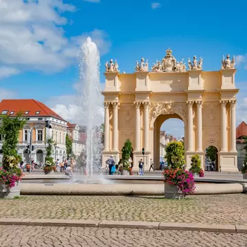 Potsdam's yellow Brandenburg Gate arch inscribed MDCCLXX, with a fountain and flower planters on a cobbled square