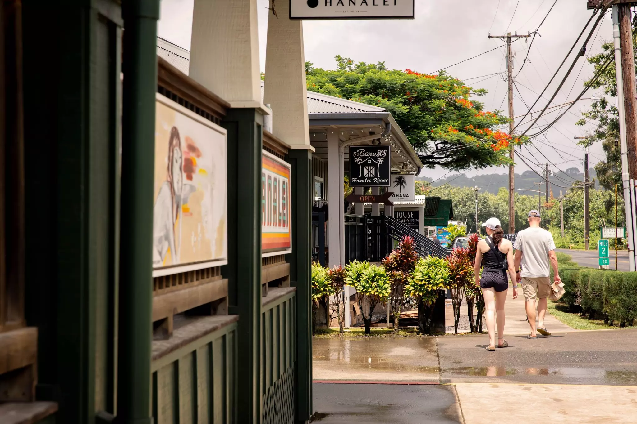 Two people walk down a sidewalk with small buildings to their left and trees in the background.