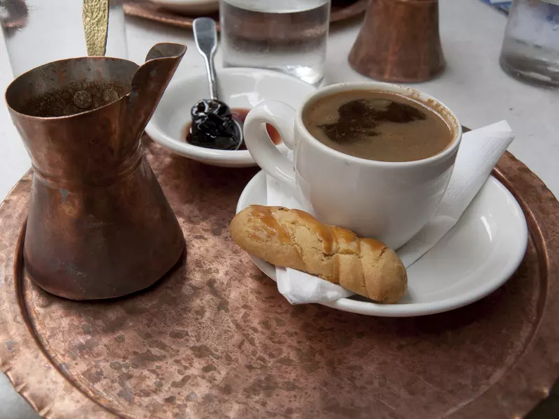 A copper tray with a white cup and saucer with coffee and a cookie, a copper vessel with coffee, and a dish with an item in dark syrup and a spoon.