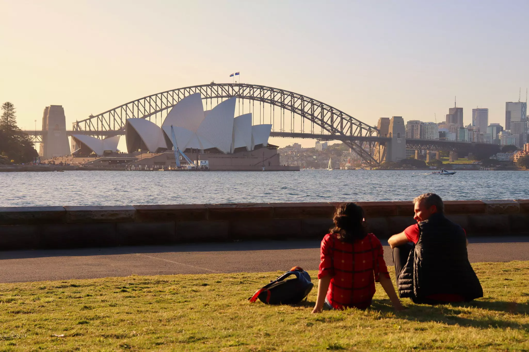Sydney Opera House and Harbour Bridge at sunset, observed by romantic couple sitting on lawn (in foreground, unfocused). Stunning view of Sydney Harbour sights.
