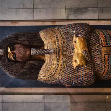 Ornate coffin with a gold face and inlaid coloured feather pattern lying in a glass display case at the Egyptian Museum