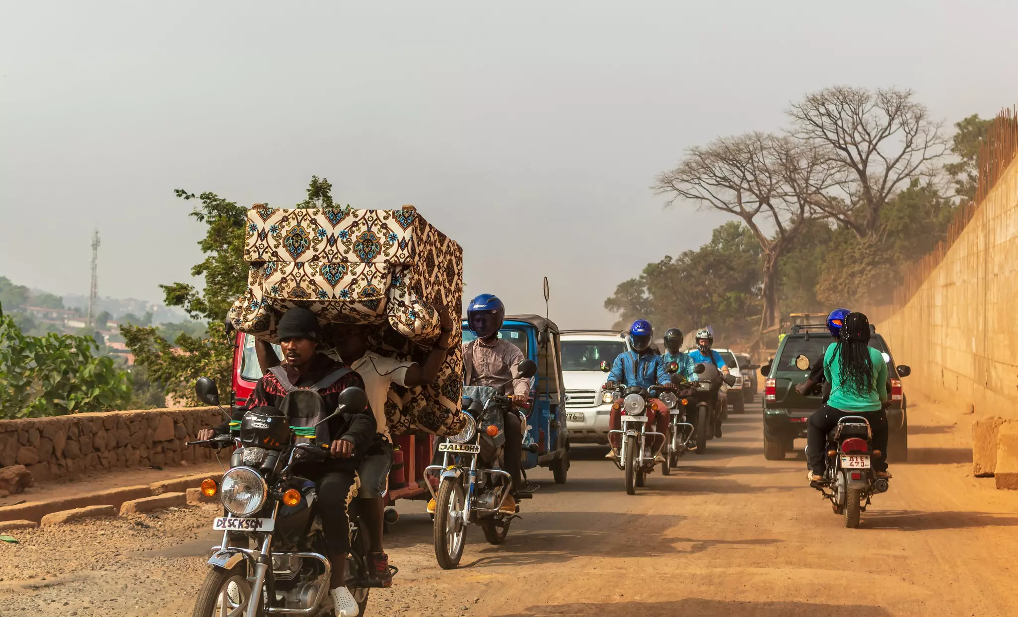 Drivers on motorcycles and in cars are pictured on a dusty road in a city.