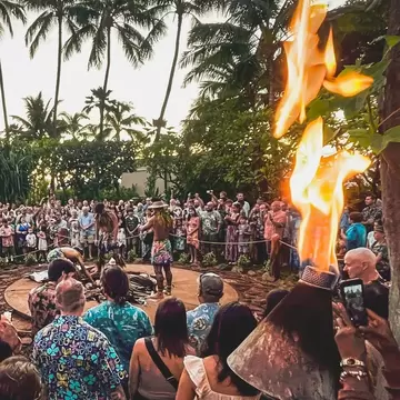 People wearing colorful patterened shirts gathered around an underground pit where people are lifting out a cooked pig: a luau in Hawaii 