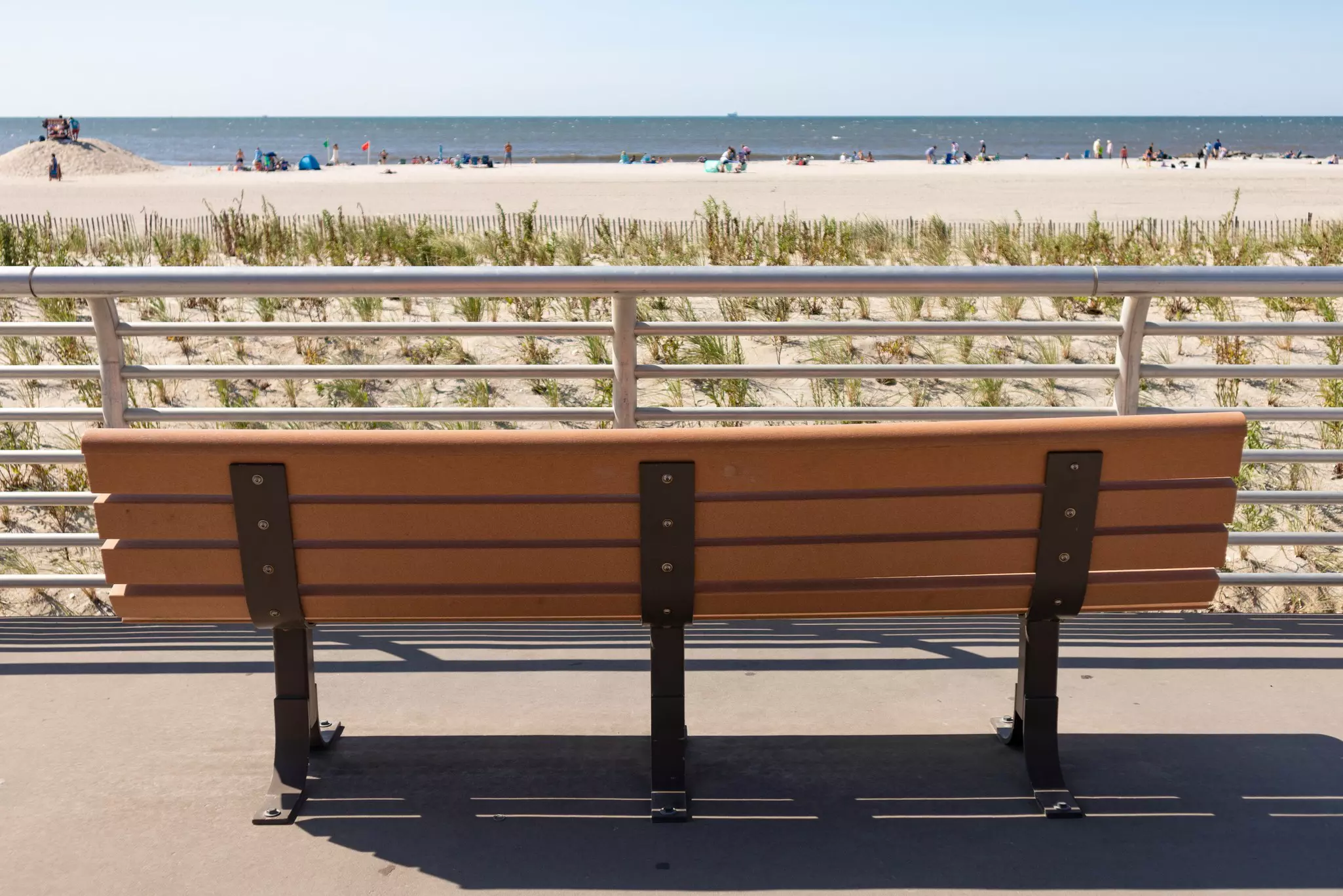 An empty bench on a boardwalk facing a sandy beach at the ocean.