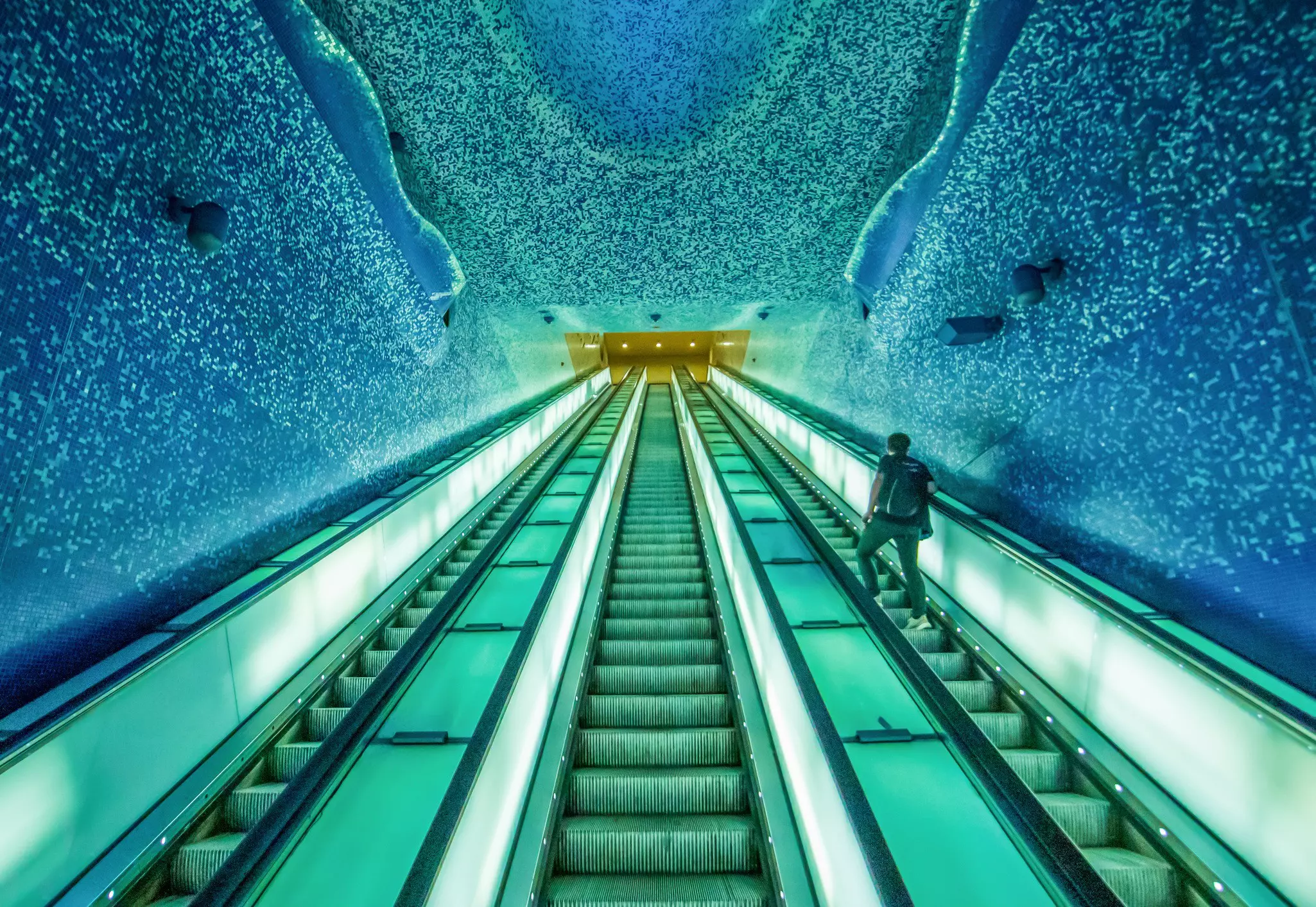 A person rides an escalator to exit a station. The space is bathed in green and blue light as part of an art installation that surrounded the stairwell.