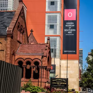 Black banner reading 'Ogden Museum of Southern Art' on an orange wall beside red-brick Confederate Memorial Hall.