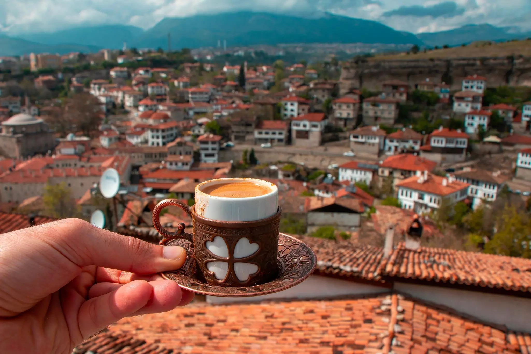 A hand holds out a small cup of coffee on a saucer with a view of a city with low-rise houses with red roofs