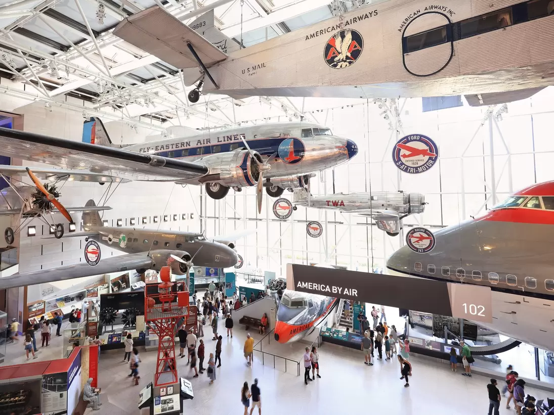 Silver propeller airliners hang from a glass-roofed hall above crowds; a black banner reads 'America by Air'.
