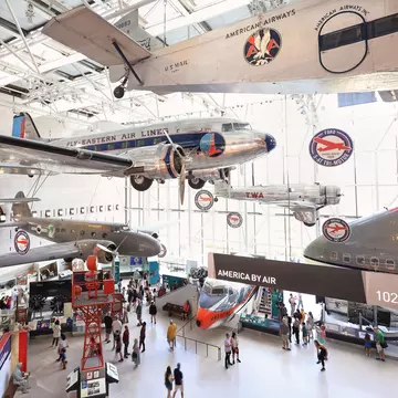 Silver propeller airliners hang from a glass-roofed hall above crowds; a black banner reads 'America by Air'.