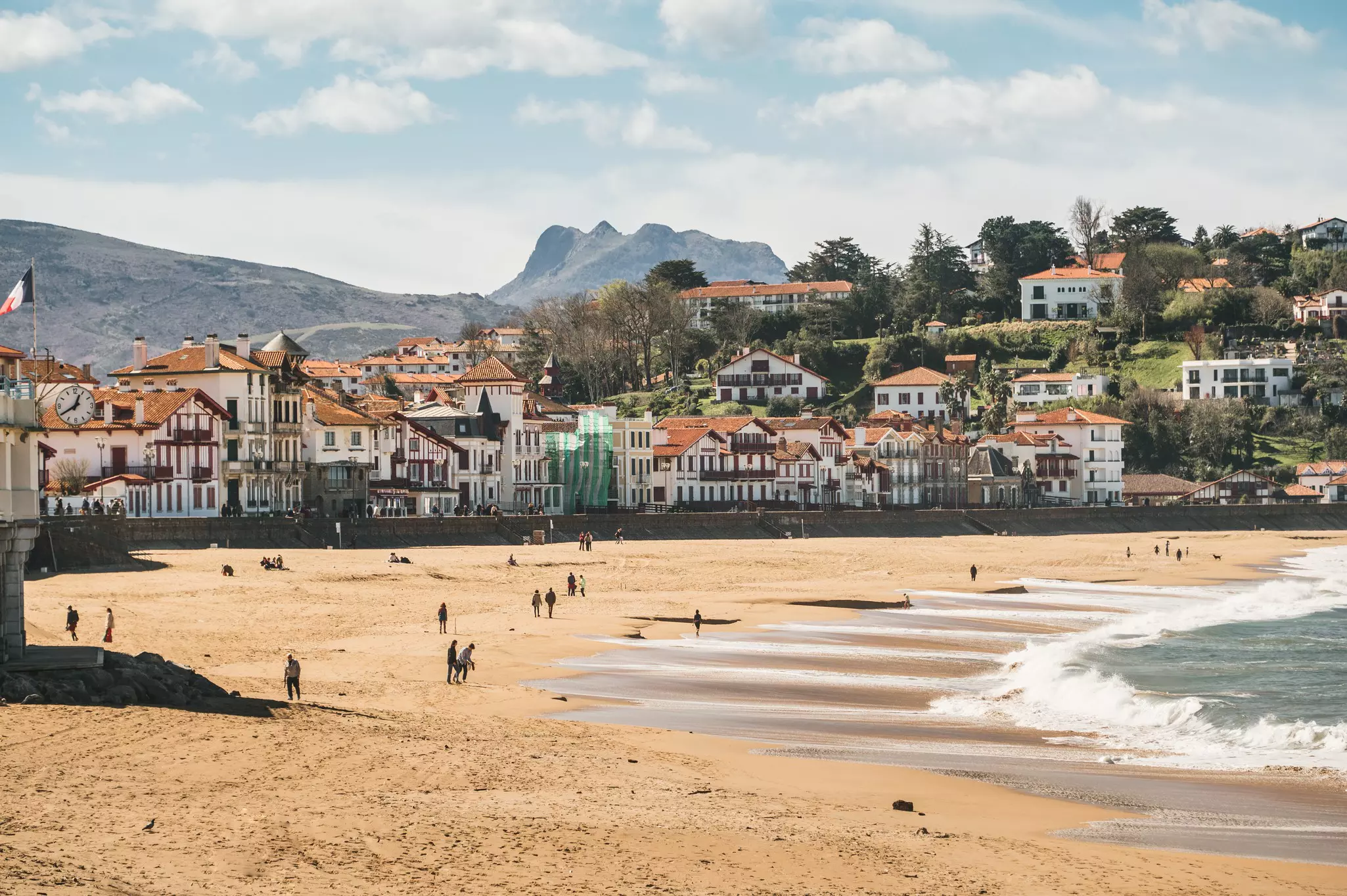 People walking along a sandy beach in a small coastal town backed by mountains.