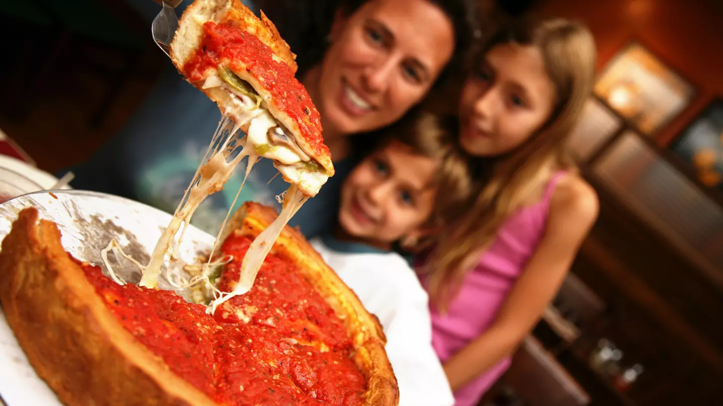 A mom lifts up a slice of Chicago deep dish pizza from a pie with spatula as two young children watch.