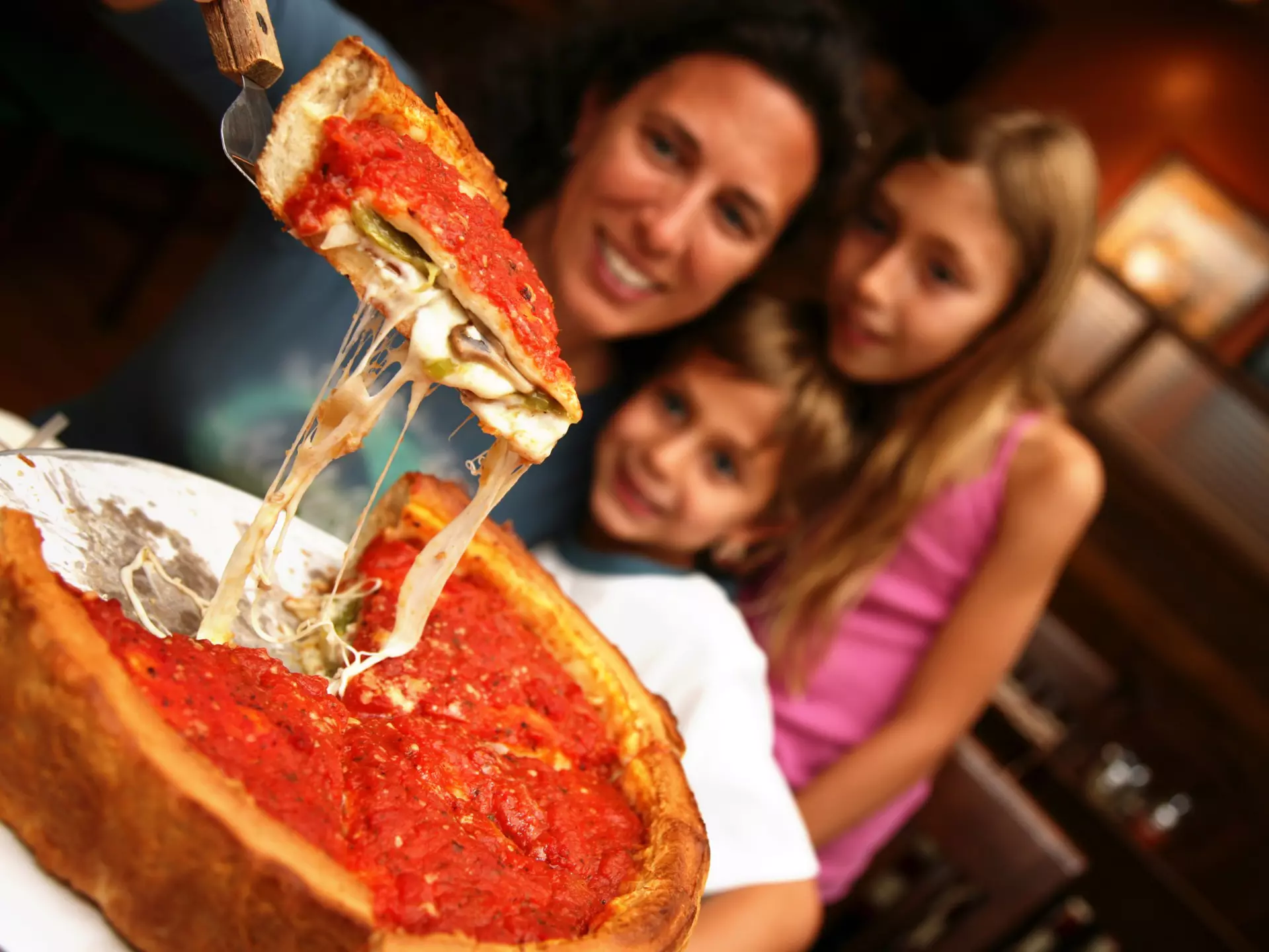 A mom lifts up a slice of Chicago deep dish pizza from a pie with spatula as two young children watch.