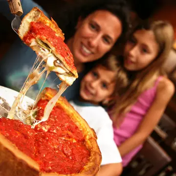 A mom lifts up a slice of Chicago deep dish pizza from a pie with spatula as two young children watch.