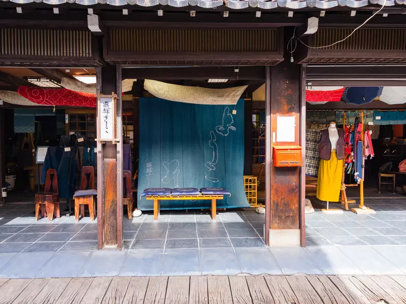 Exterior of fabric shop with a large indigo-dyed cloth in the center and a mannequin with long yellow skirt and brown vest to the right.