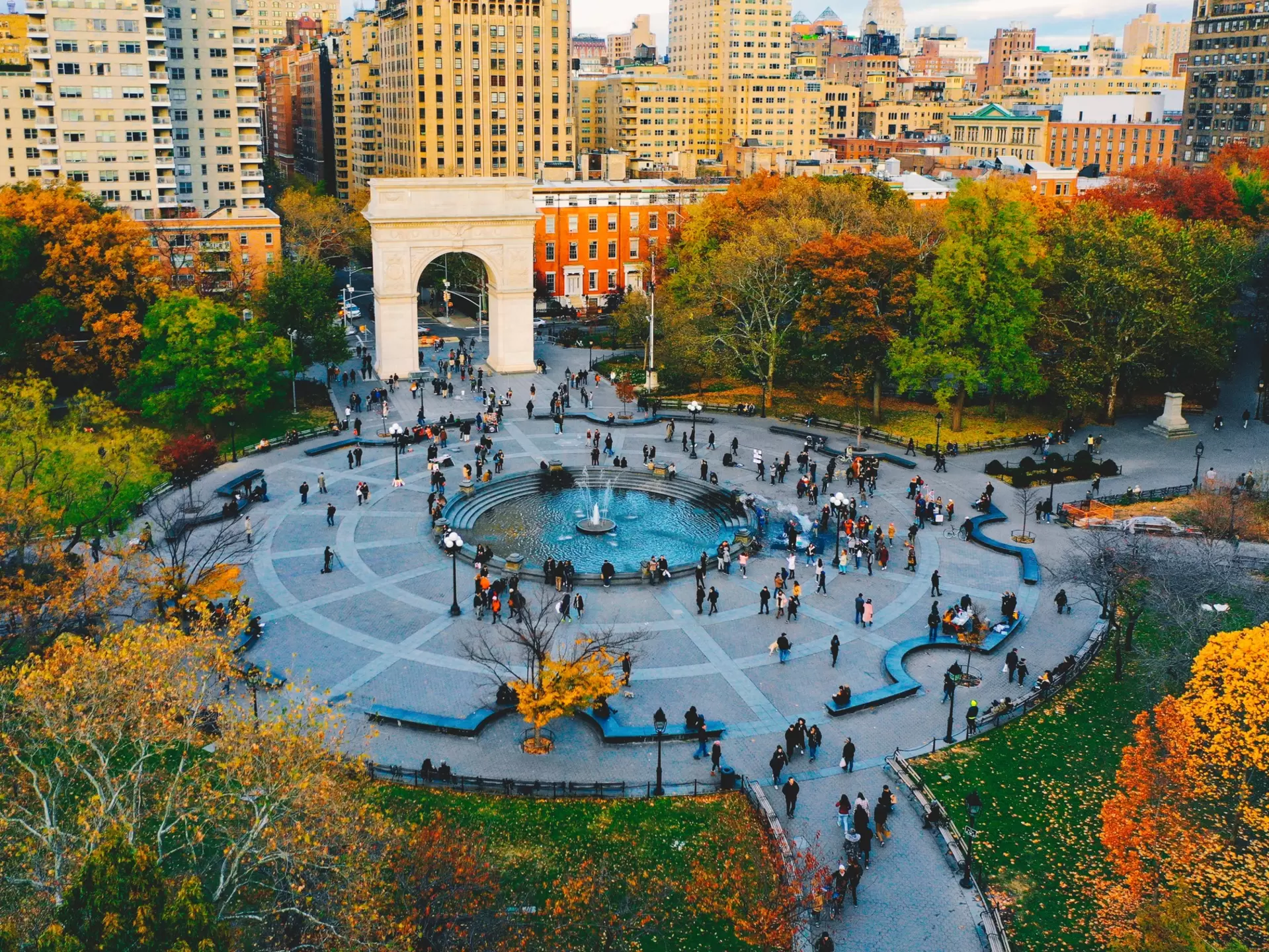 People milling about a circular plaza with a fountain at its center in New York City. Fall foliage surrounds the plaza.