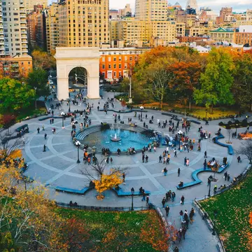 People milling about a circular plaza with a fountain at its center in New York City. Fall foliage surrounds the plaza.
