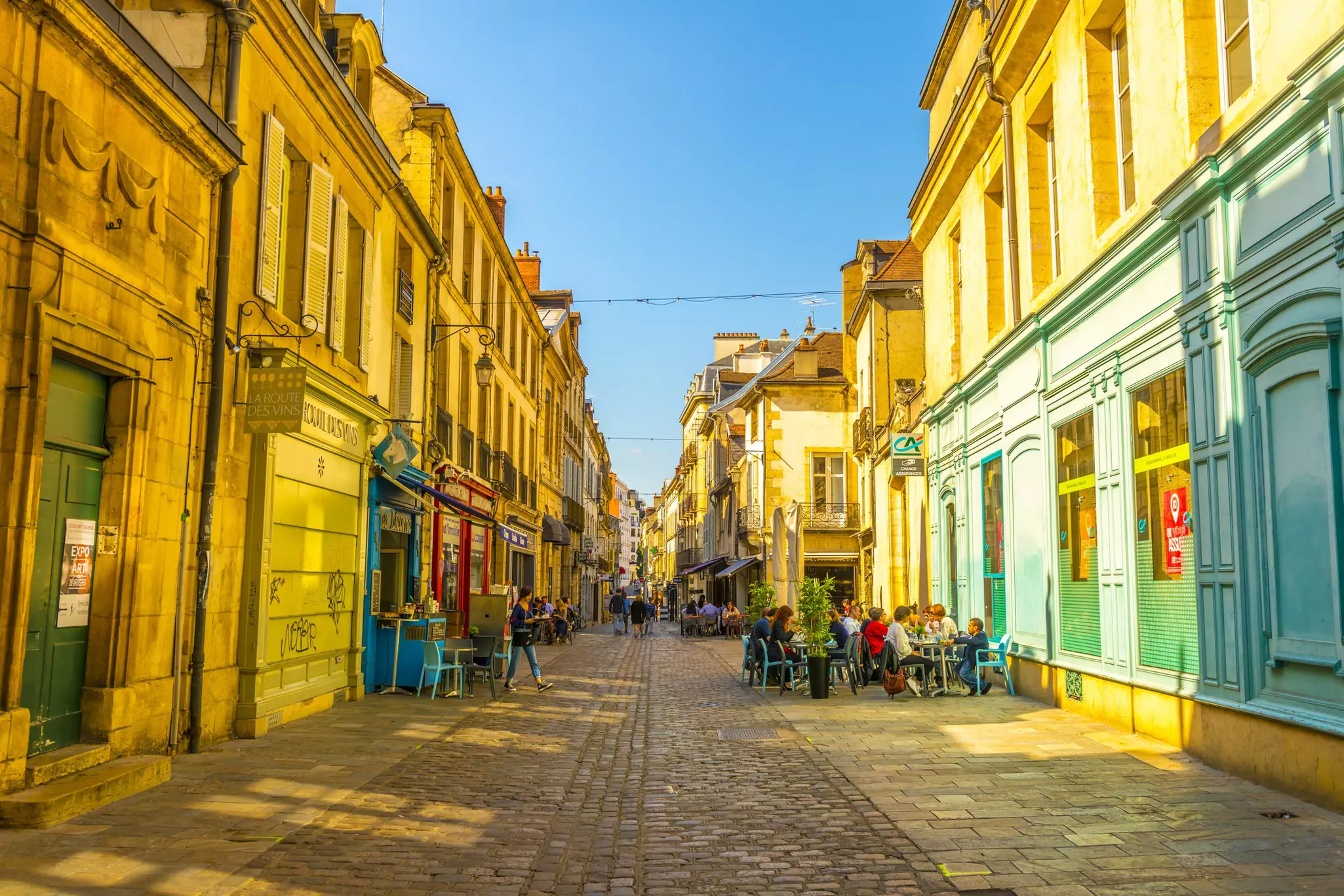 Sunset on a cobbled street with tables and chairs outside colorful cafes and people eating and drinking