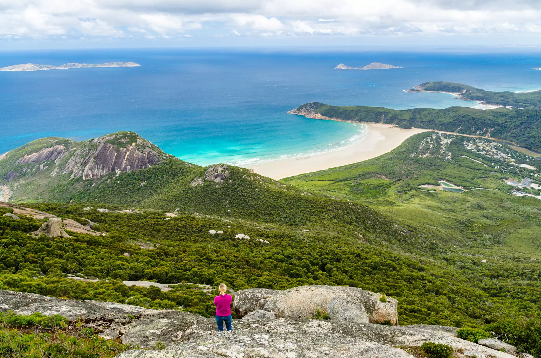 Aerial viwe of solo woman on rocky hillside overlooking greenery and the ocean beyond.