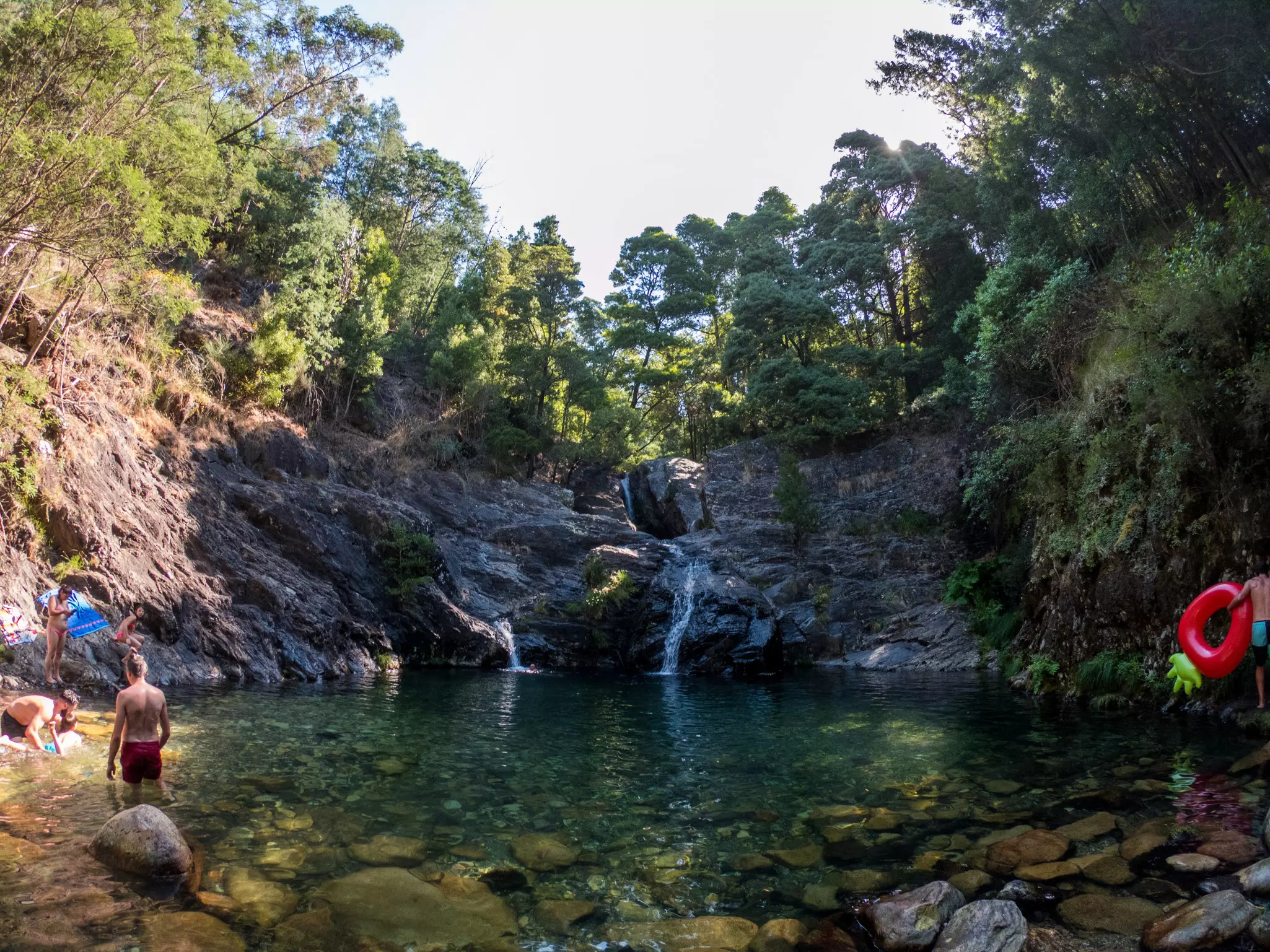 People swim in the lagoon formed by a waterfall, shaded by trees.