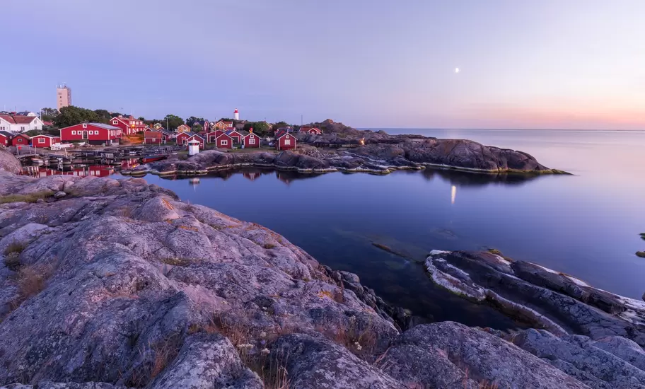 Red wooden houses and a lighthouse on smooth granite rocks by calm sea at dusk in Sweden's coastal archipelago