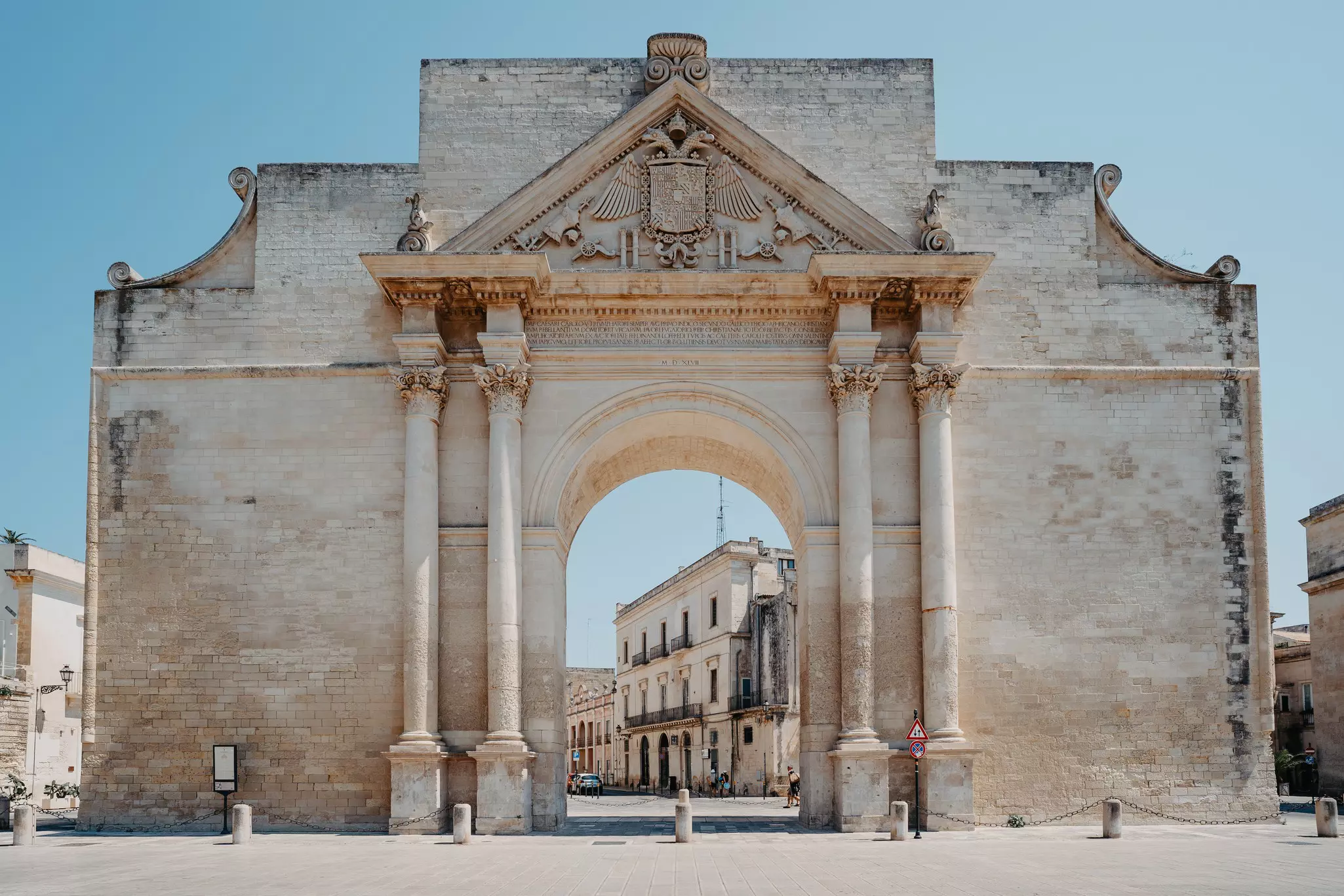 A huge baroque archway stands in a city square, framing a view of stone buildings on a street behind.