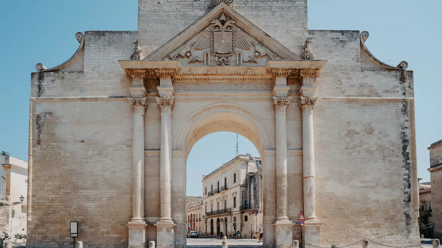 A huge baroque archway stands in a city square, framing a view of stone buildings on a street behind.