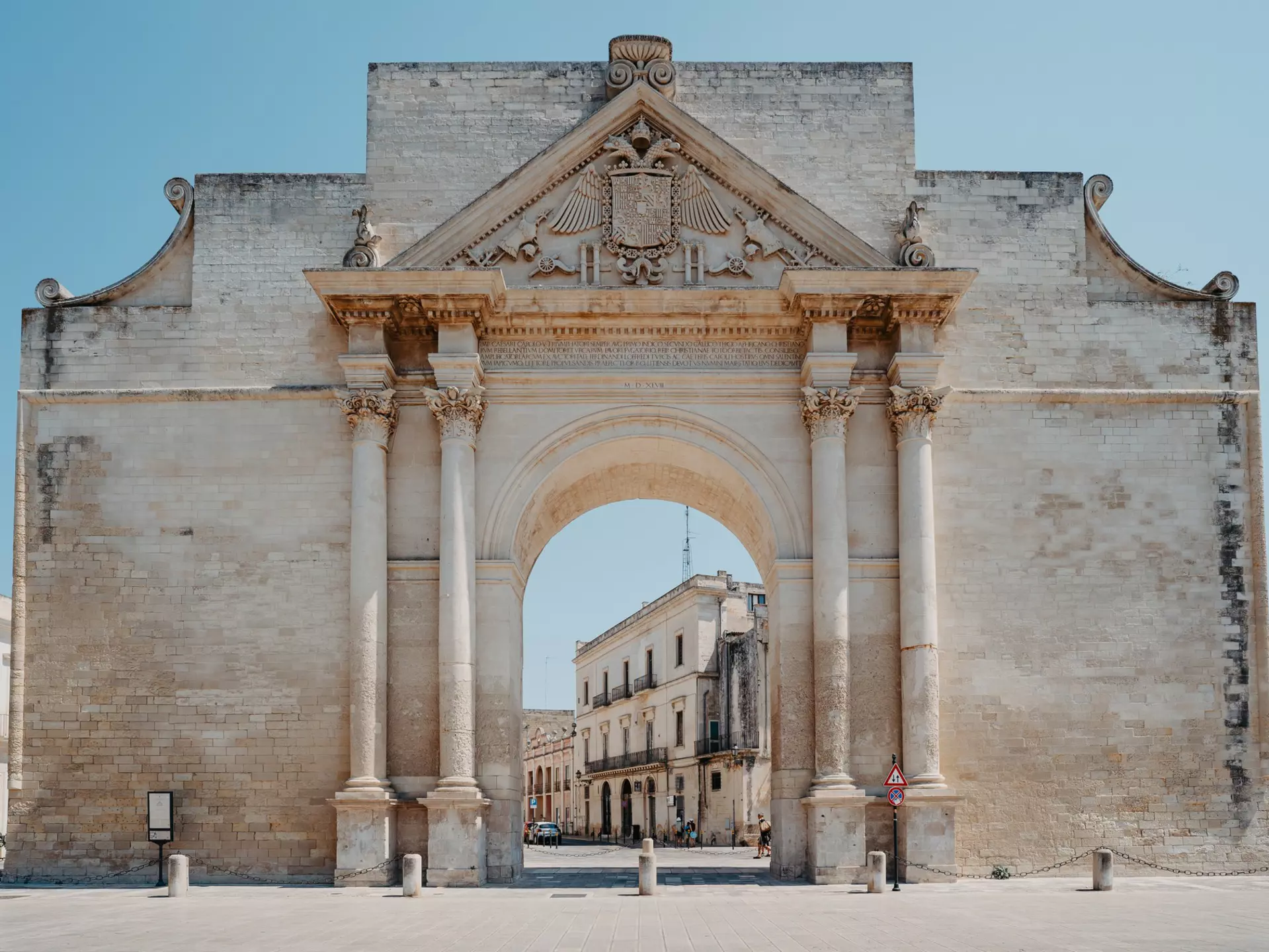 A huge baroque archway stands in a city square, framing a view of stone buildings on a street behind.