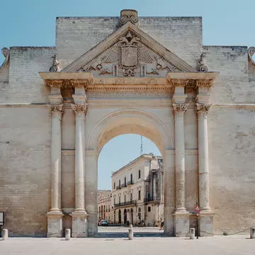 A huge baroque archway stands in a city square, framing a view of stone buildings on a street behind.