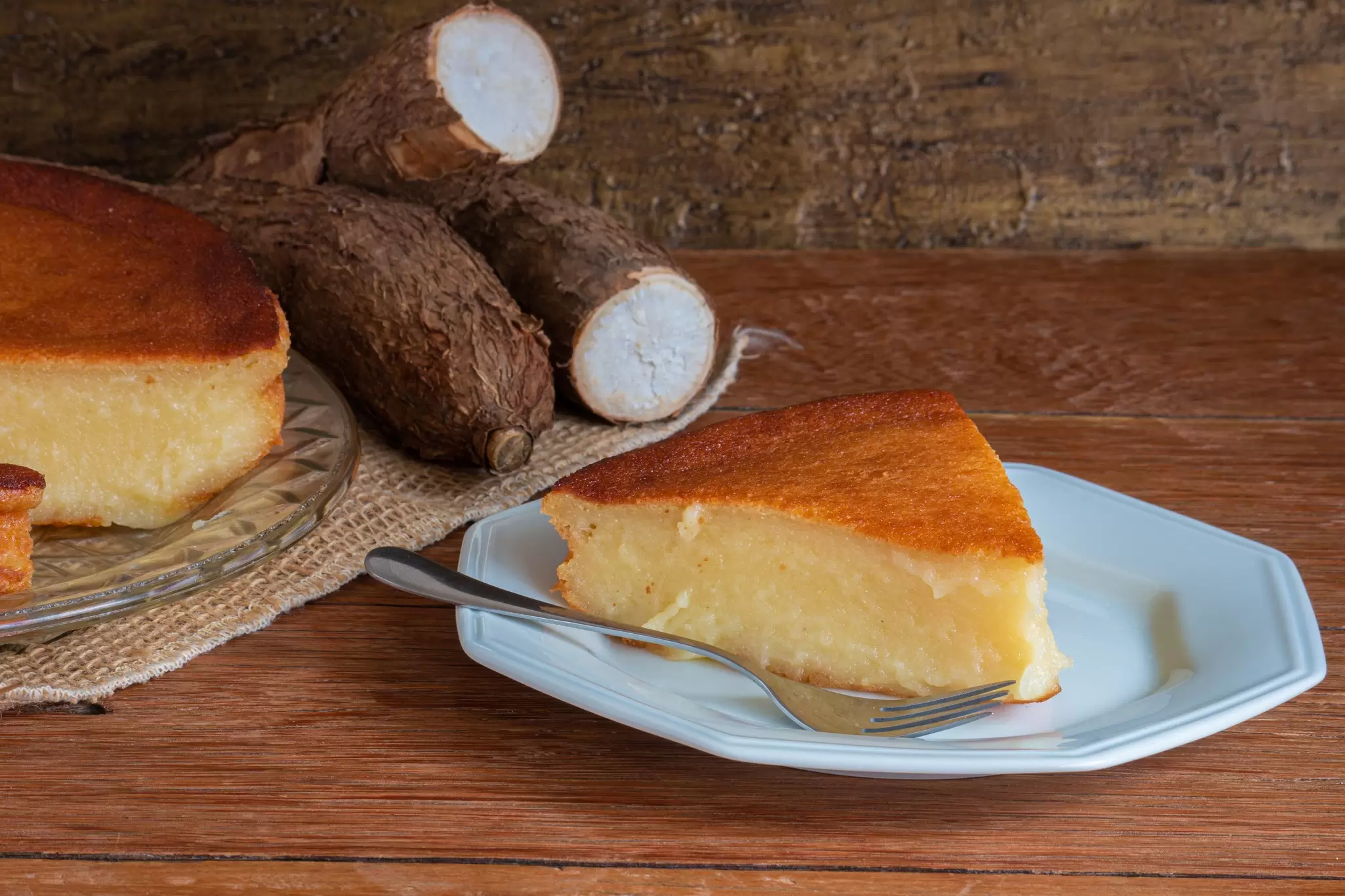 A slice of dense cassava pie on a plate next to the full pie and cassava roots