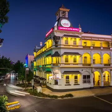 Ornate three-storey heritage hotel with arched verandas glowing yellow at dusk on a street corner, car light trails passing.