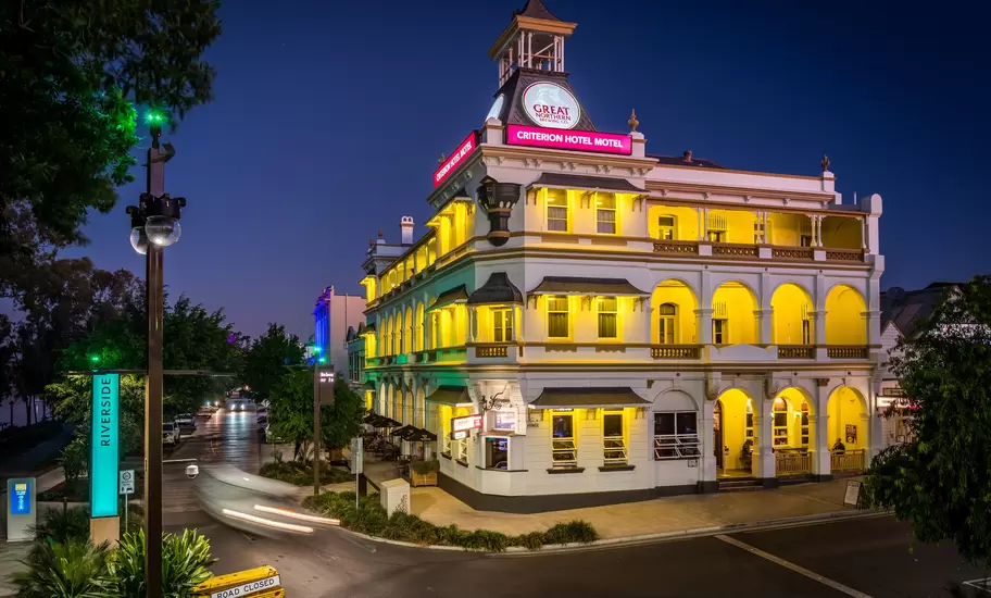 Ornate three-storey heritage hotel with arched verandas glowing yellow at dusk on a street corner, car light trails passing.