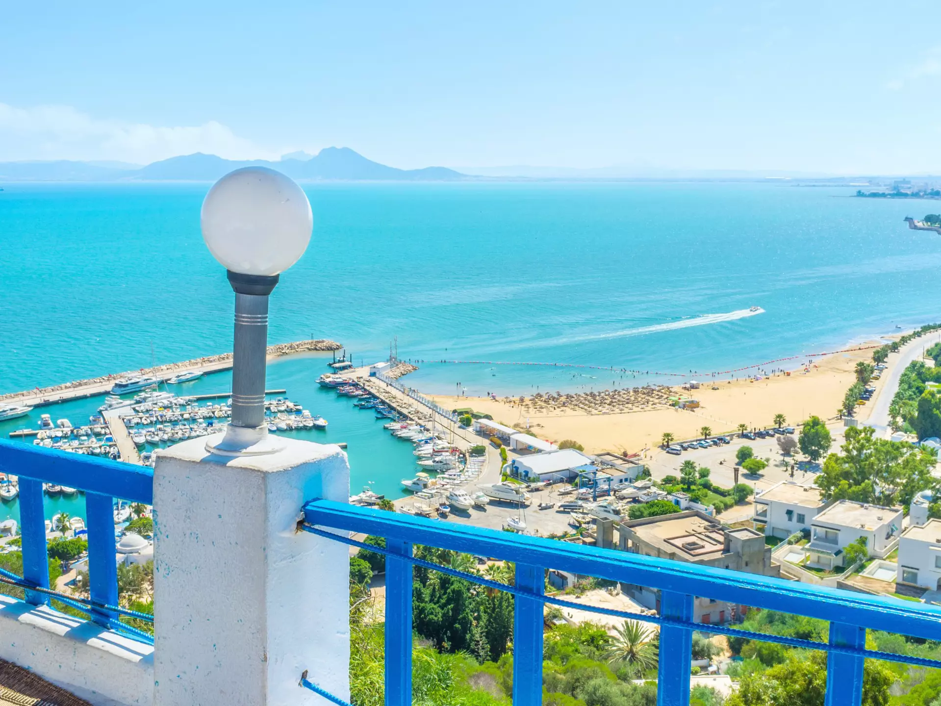 A view from a terrace (protected by a blue-painted parapet) overlooking a marina and small beach in a town.
