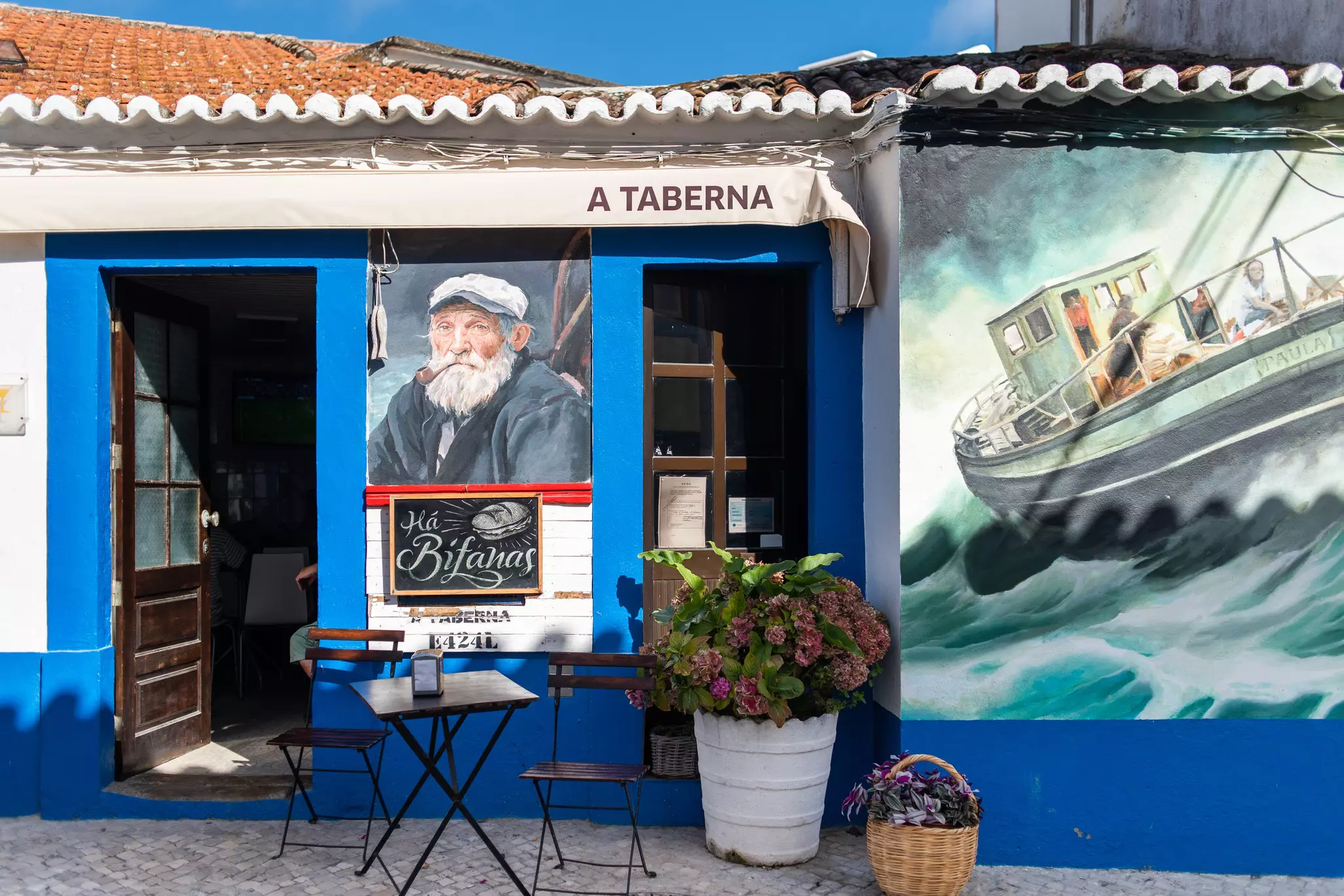 The exterior of a small taverna. The outside has a table, chairs and flower pots with a giant wall mural of a fishing boat and a smaller mural of a fisherman with a pipe in his mouth.