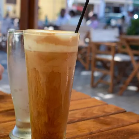 A tall clear glass with a coffee drink with foam on top on a wooden table.