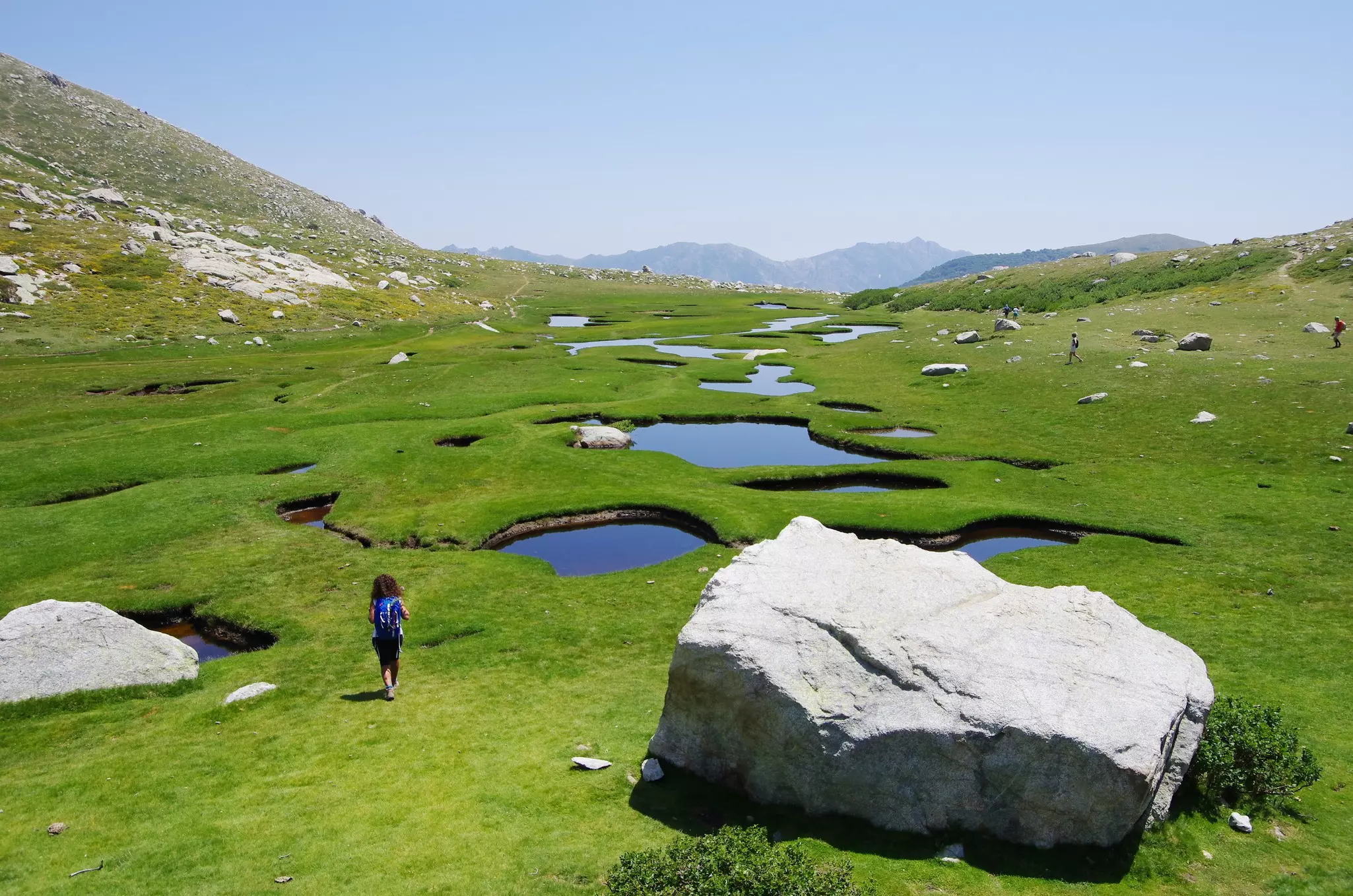 A woman walks across a lush grassland dotted with small ponds heading towards mountains.