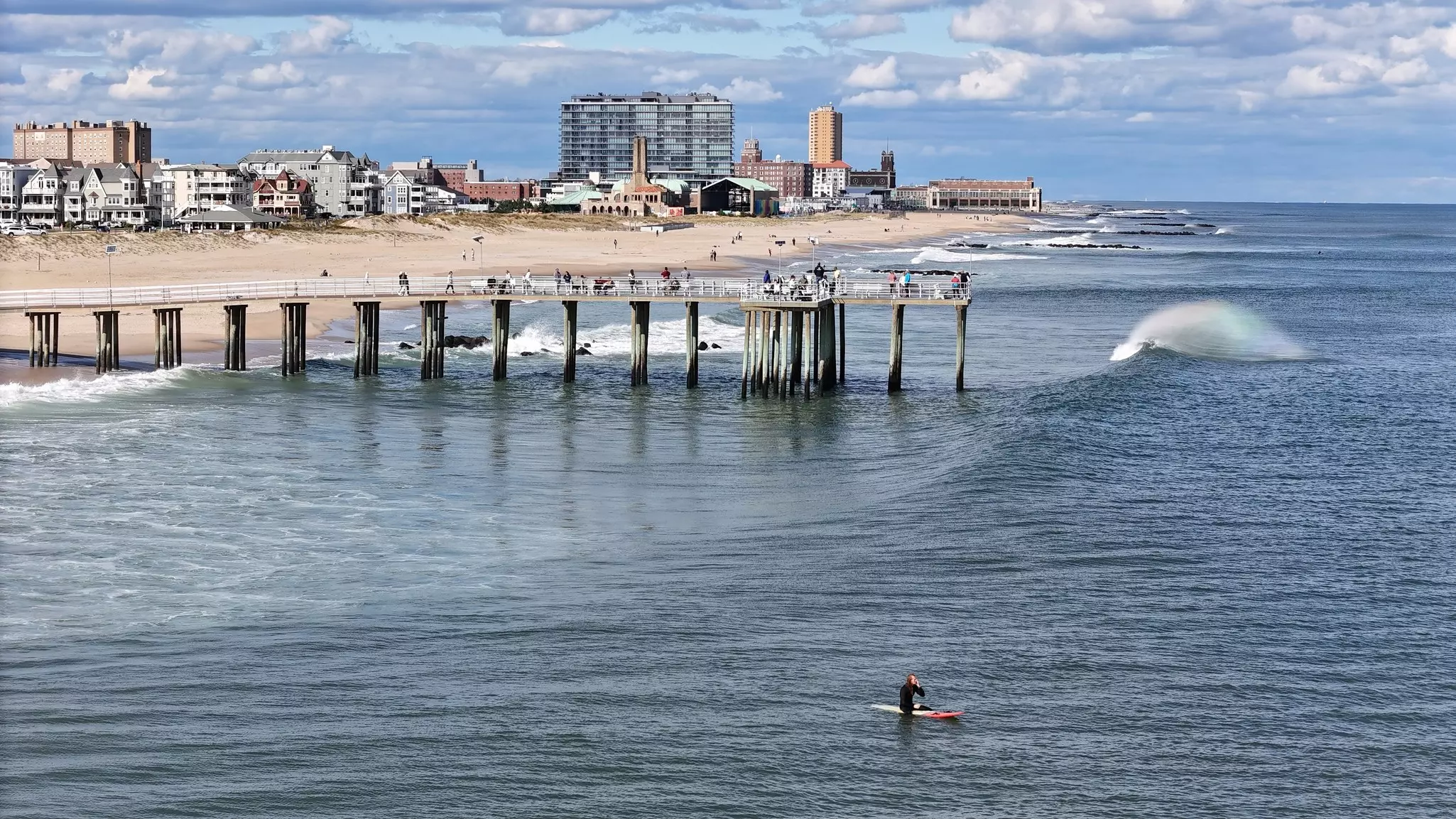 A surfer sits on a board offshore from a beach town with a small pier.