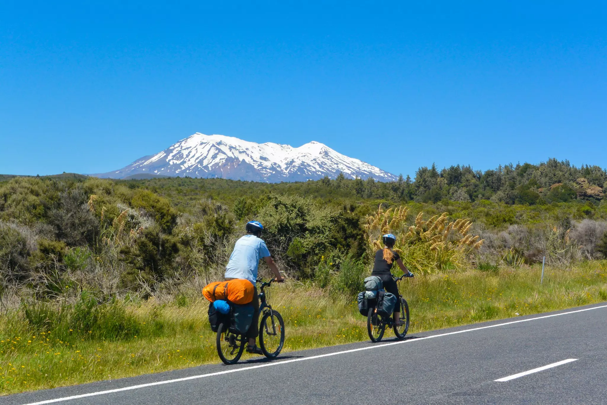 Two cyclists with fully loaded bikes pedal along a road within sight of a snow-capped mountain.