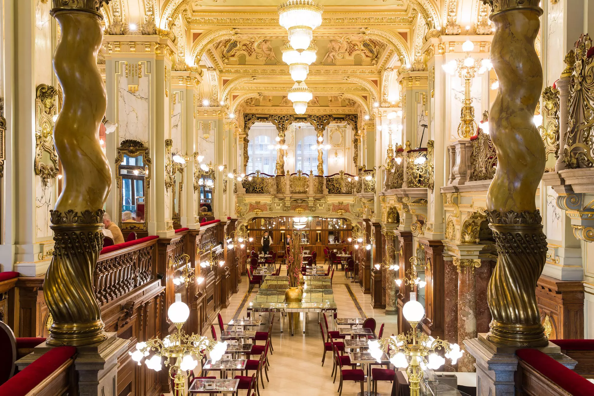 The luxurious interior of a cafe in Budapest; there are marble columns, golden light fixtures and plush crimson chairs.