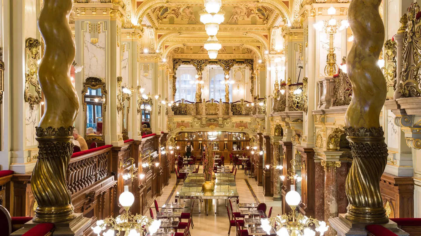 The luxurious interior of a cafe in Budapest; there are marble columns, golden light fixtures and plush crimson chairs.