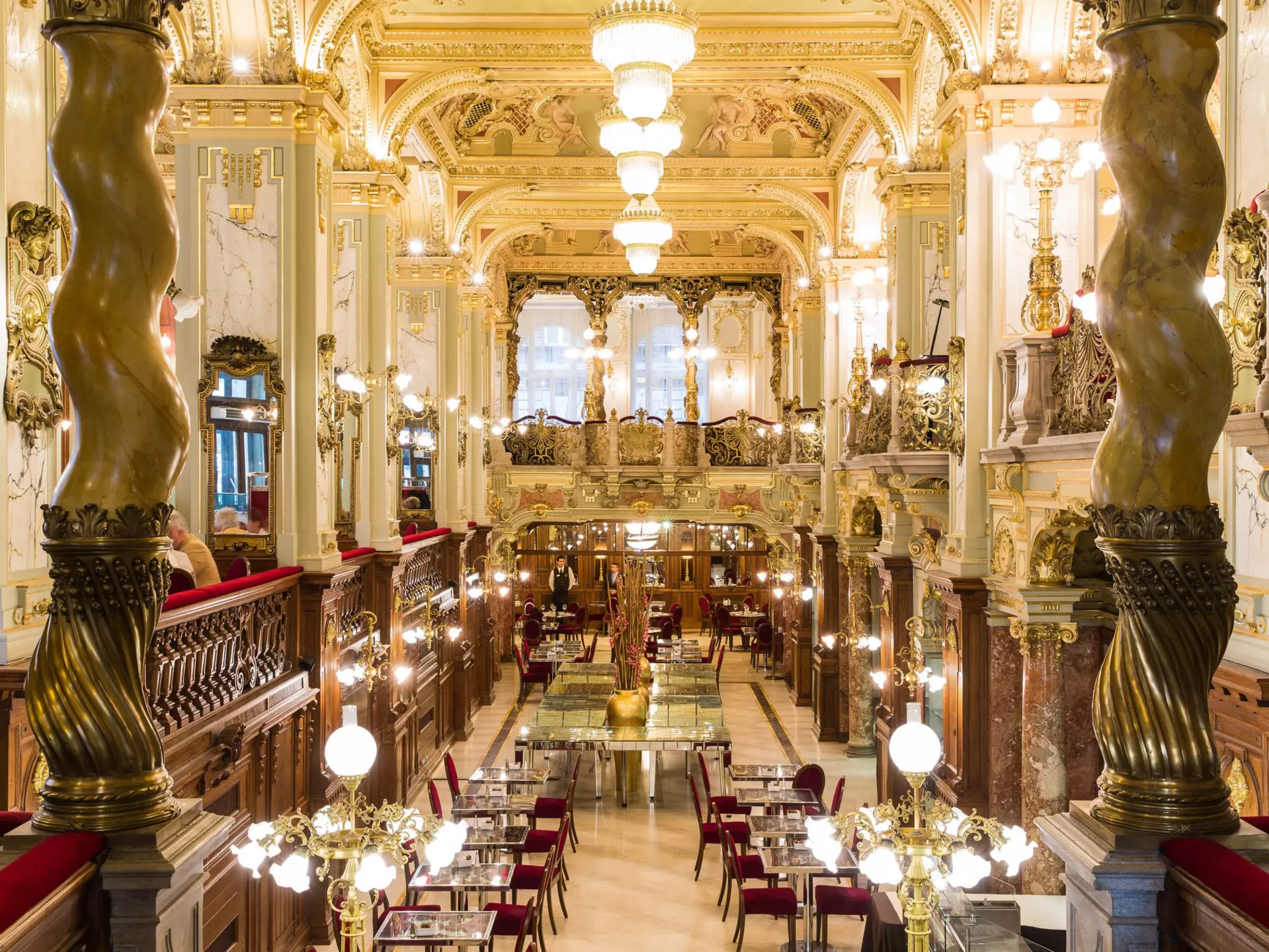 The luxurious interior of a cafe in Budapest; there are marble columns, golden light fixtures and plush crimson chairs.