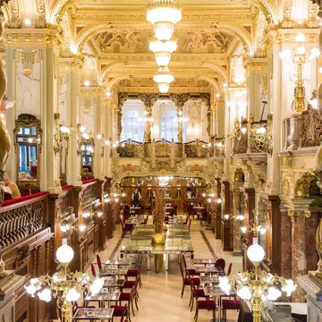The luxurious interior of a cafe in Budapest; there are marble columns, golden light fixtures and plush crimson chairs.