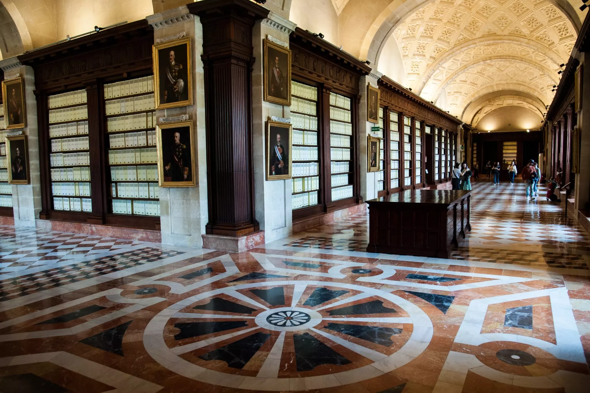The hallways of an elaborate builing have inlaid marble tiles on the floor, barrel-vaulted ceilings, and hundreds of bookshelves.
