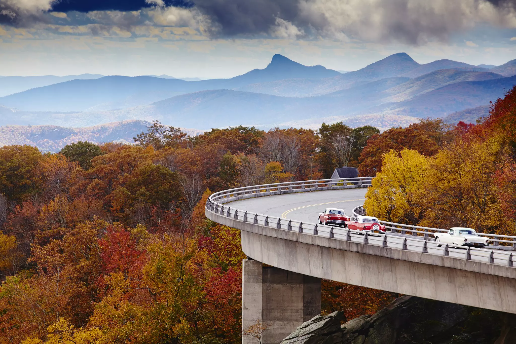 Vintage cars drive along a curved road built above a hillside covered with trees showing fall foliage. Hazy mountains are visible in the distance.
